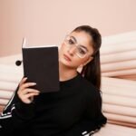 A young woman in sportswear and eyeglasses reads a book indoors, exuding focus and style.
